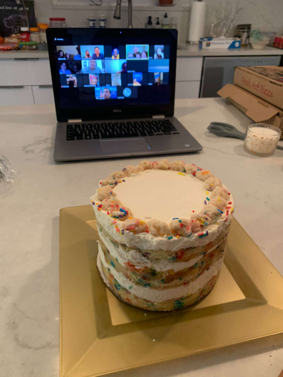 A funfetti layer cake sits on a square gold plate on a kitchen counter. Behind it, a laptop displays a virtual group video call, and a pizza box and candle are nearby.