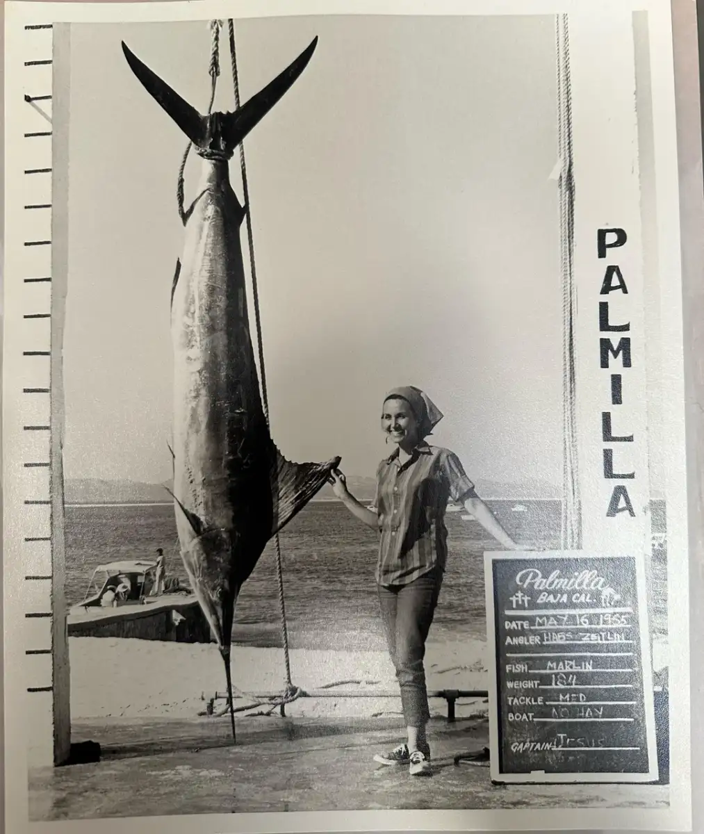 A woman poses next to a large marlin hanging vertically on a dock, with a sign listing fishing details and the word "PALMILLA" on a post; the sea and a small boat are visible in the background.