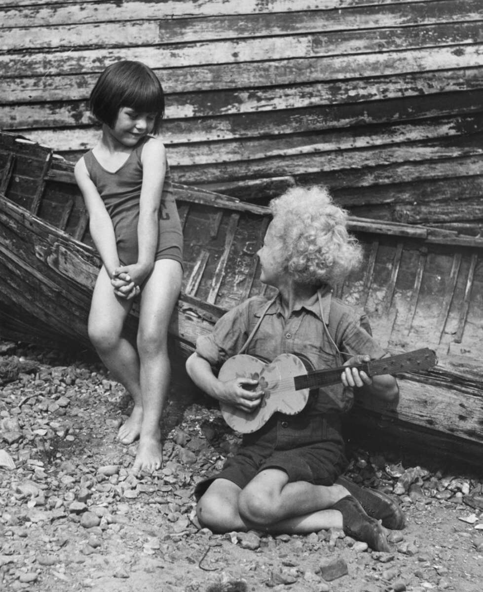 A barefoot boy with curly blond hair plays a small banjo while kneeling on rocky ground, smiling up at a barefoot girl in a swimsuit who stands nearby in front of an old wooden boat.