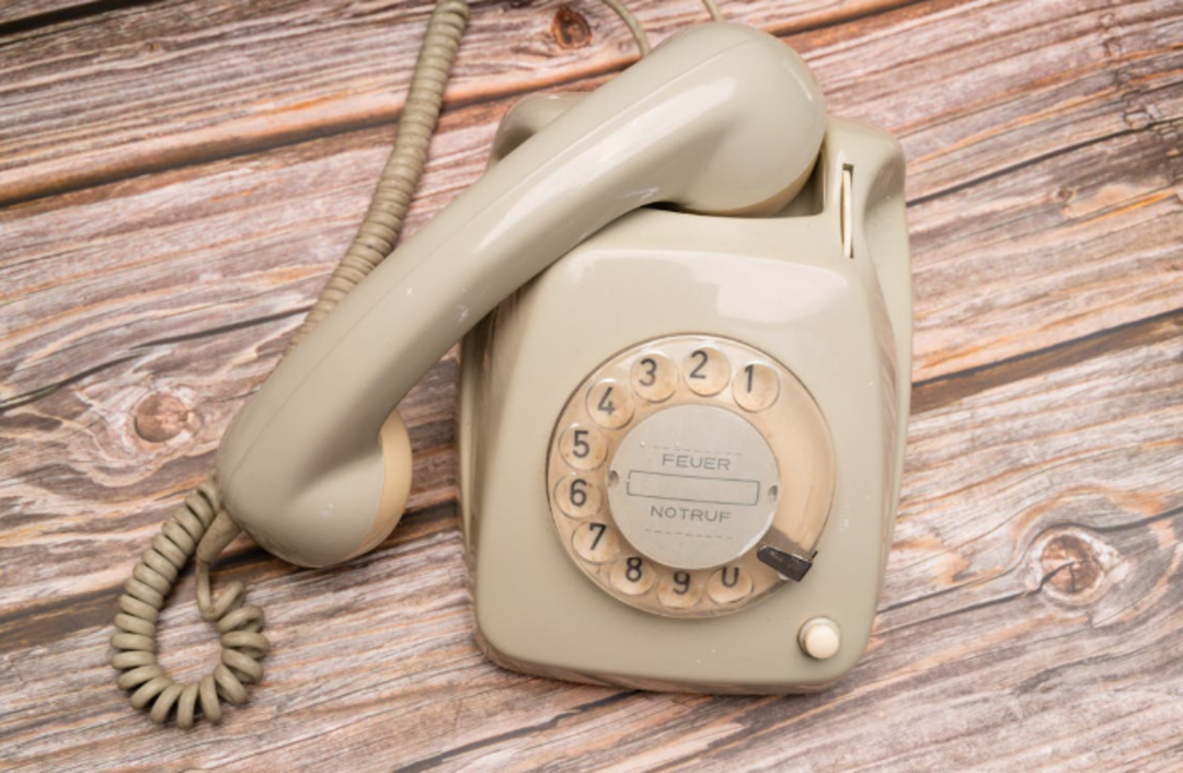 A beige rotary dial telephone with a coiled cord sits on a wooden surface. The dial has numbers and German words "FEUER" and "NOTRUF" visible in the center.