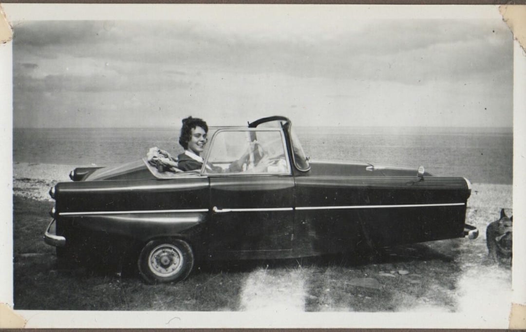 A person smiling while sitting in a convertible parked by the seaside, with the ocean and cloudy sky in the background. The photo is in black and white.