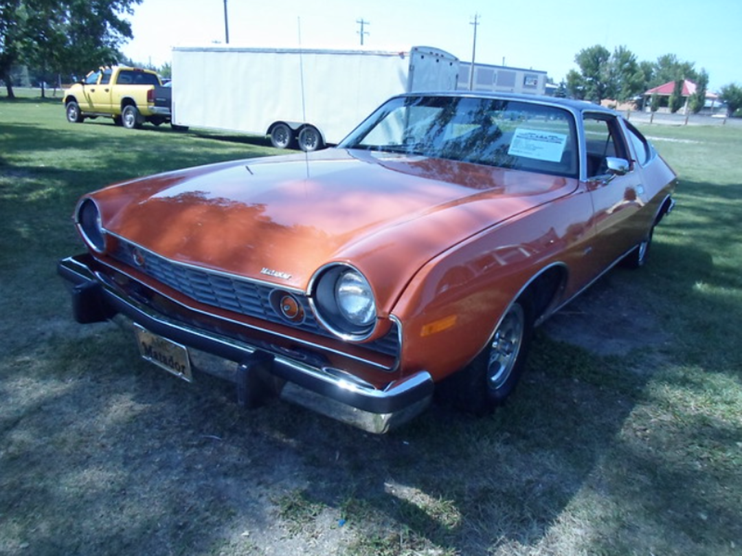An orange vintage Ford Pinto car is parked on grass near a white trailer and a yellow truck, with trees and buildings visible in the background.