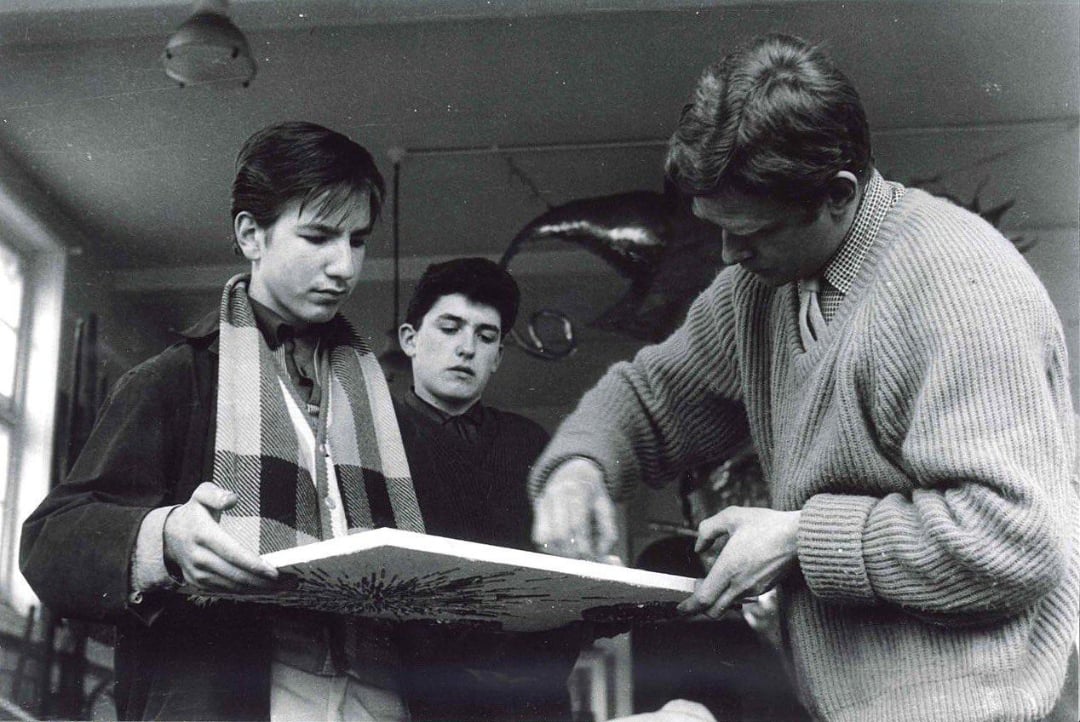 Three people stand indoors; one adult on the right gestures over a flat object held by a boy in a scarf, while another boy stands behind them, watching attentively. The scene appears to be instructional.