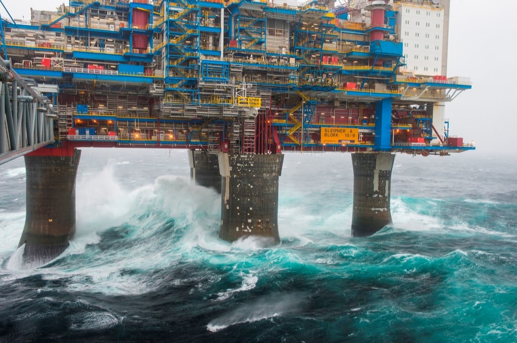 A large offshore oil platform stands above rough, choppy ocean waves, with water crashing against its massive support pillars under a cloudy, overcast sky.