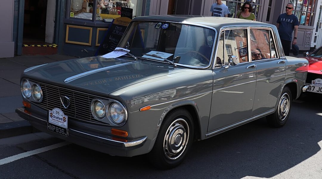 A vintage grey classic car with chrome detailing is parked on a street in daylight, with a paper on its windshield. Several people and storefronts are visible in the background.