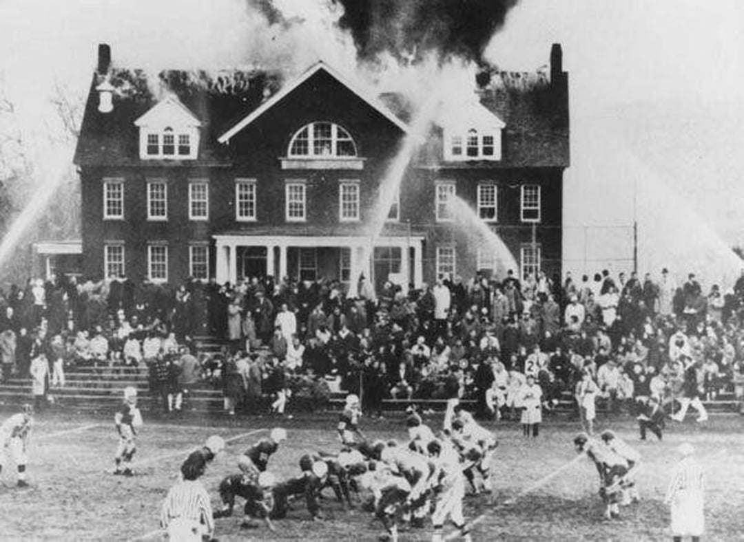 A football game is being played while a large crowd watches a building in the background with its roof on fire, as firefighters spray water to extinguish the flames.