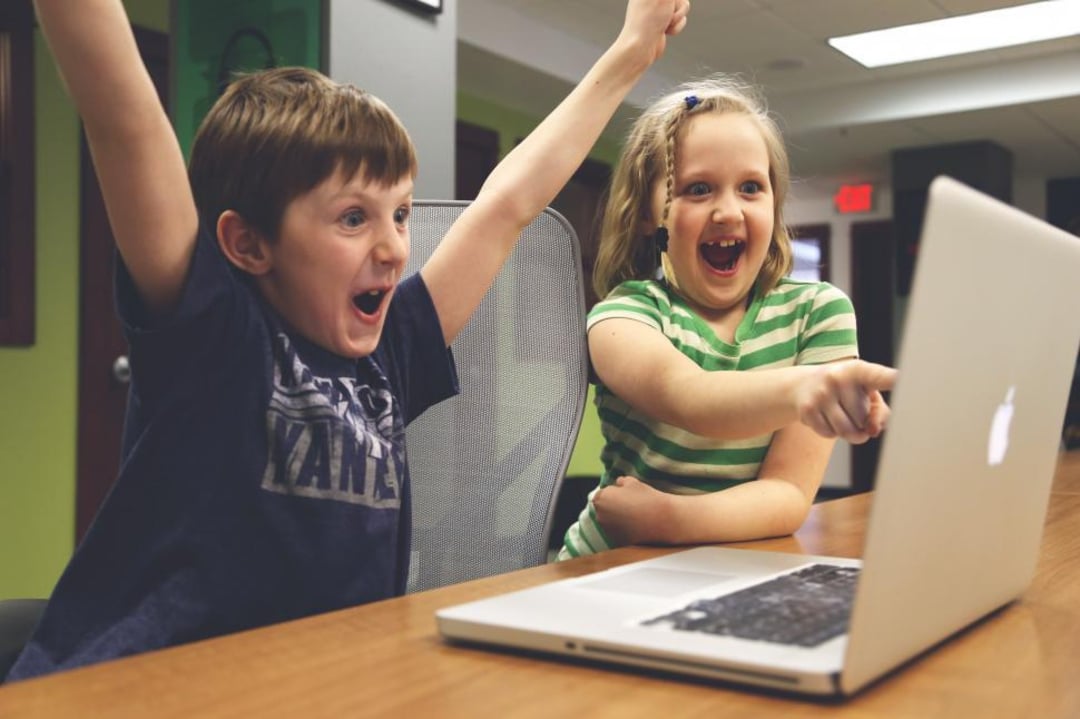 Two excited children sit at a table, cheering and smiling while looking at a laptop screen; one child has arms raised in excitement, the other points at the screen.