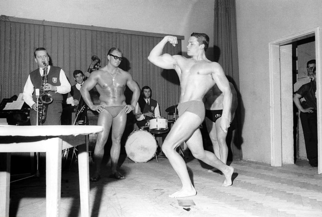 A young male bodybuilder in swim trunks flexes his arm on stage while another bodybuilder and several musicians, including a saxophonist and drummer, look on in a black-and-white photo.