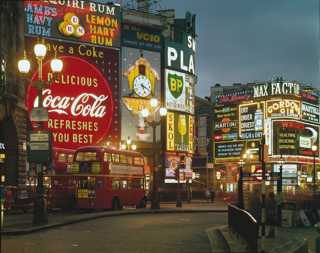 Bright neon advertising signs light up a city square at night, featuring brands like Coca-Cola and BP. A classic red double-decker bus is parked on the street, and old-fashioned street lamps glow nearby.