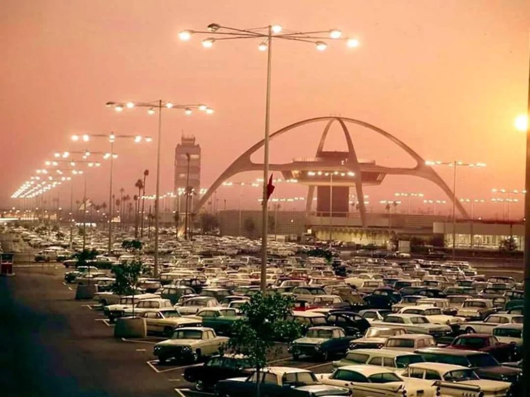 A large parking lot filled with vintage cars at sunset, with the iconic Theme Building and control tower of Los Angeles International Airport (LAX) in the background under glowing streetlights.