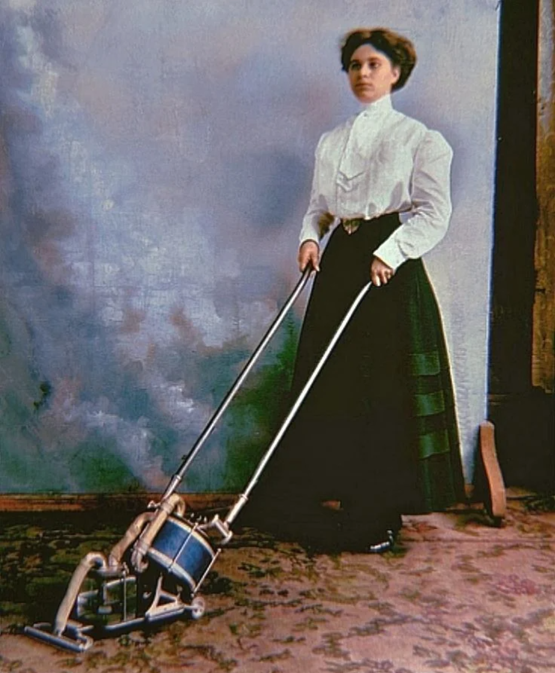 A woman in early 20th-century clothing stands upright on a patterned carpet, holding the handles of a vintage vacuum cleaner with metal rods and a blue canister. The background is neutral with muted tones.
