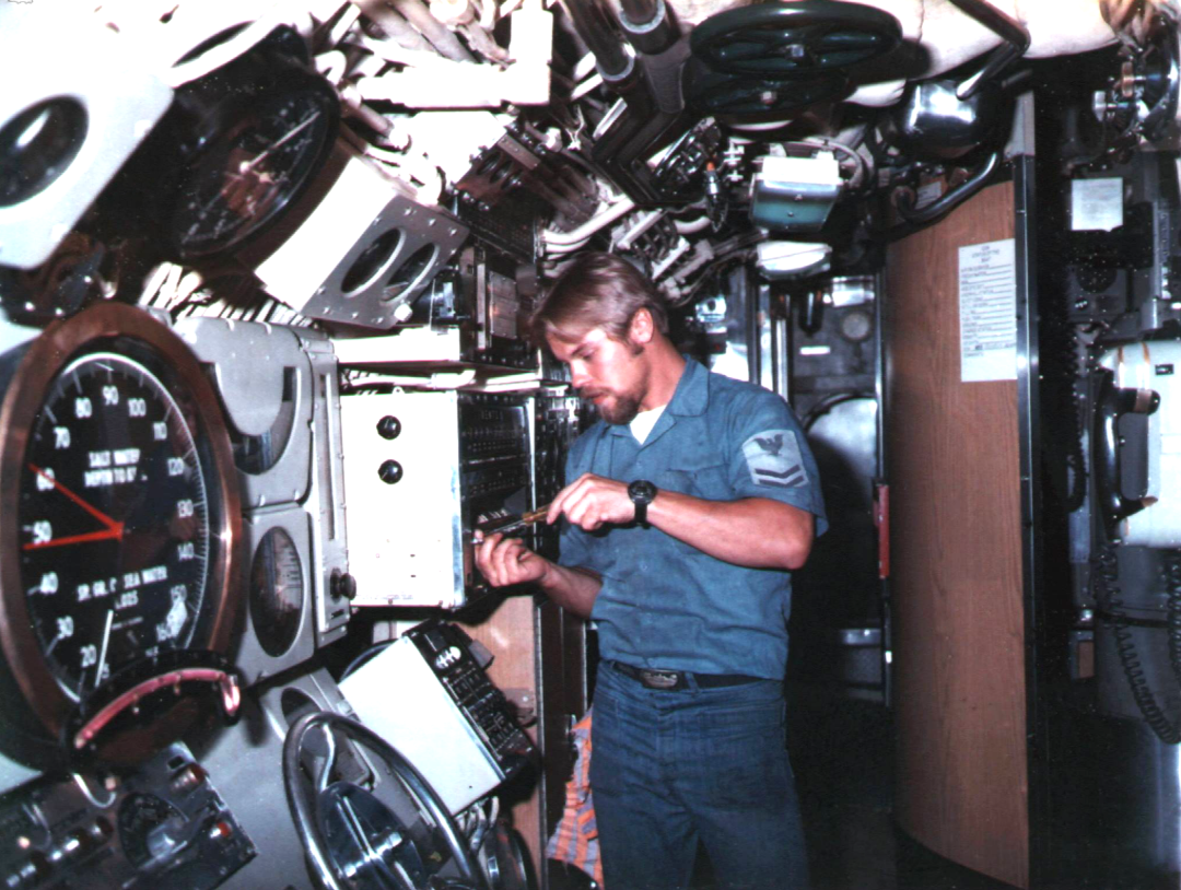A man in a blue uniform uses tools to work on electrical equipment inside a submarine, surrounded by dials, gauges, and machinery.