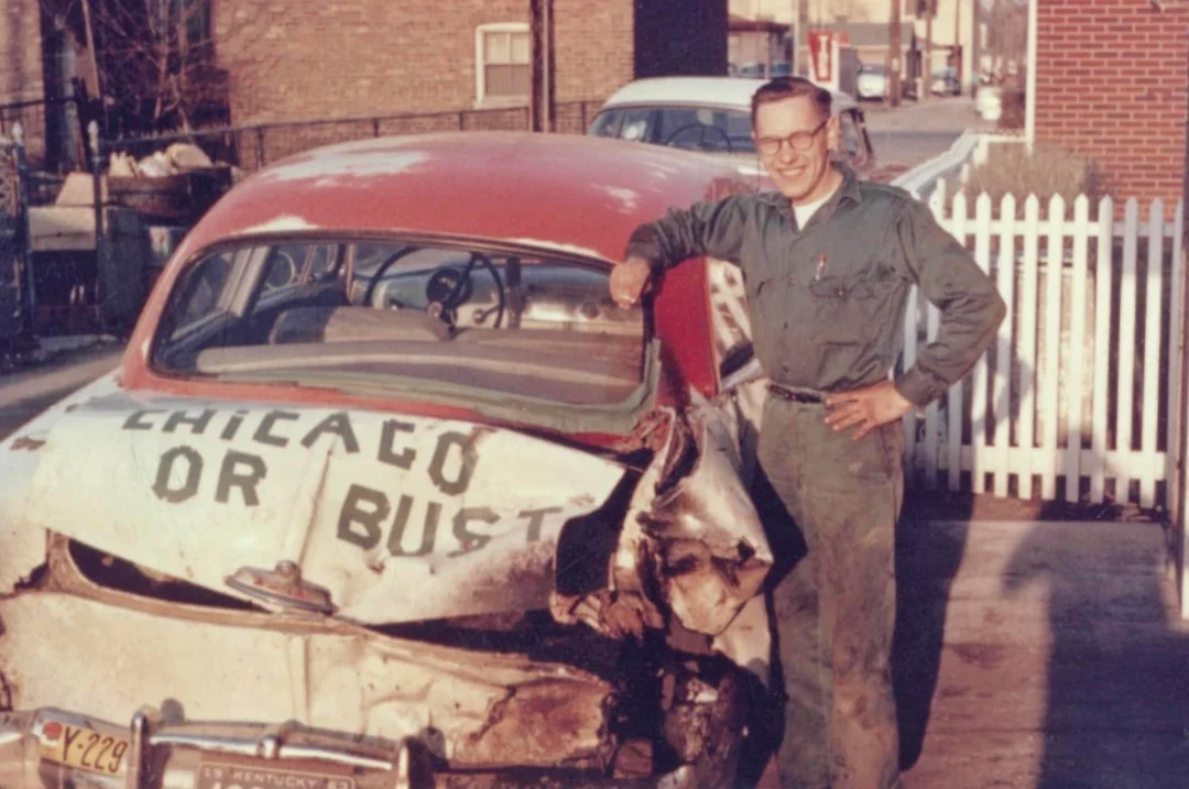 A man in glasses and a jumpsuit smiles and poses beside a vintage car with a damaged rear end. The car's trunk is painted with the words "CHICAGO OR BUST." The photo appears to be from the mid-20th century.