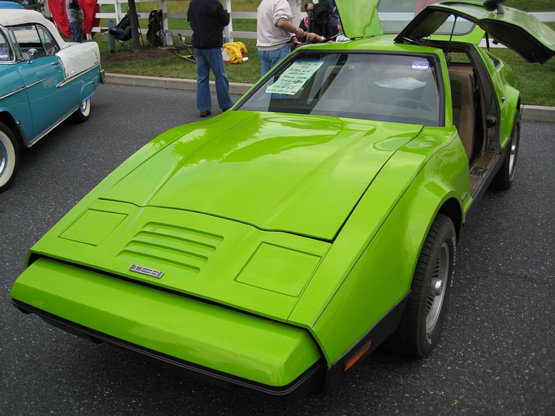 A bright green vintage sports car with gull-wing doors open, parked at a car show. Several people stand nearby, and another classic car is visible in the background.