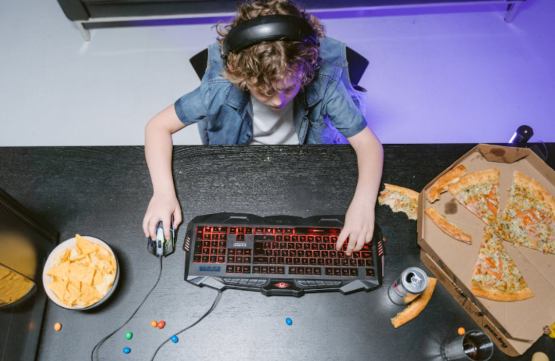 A child wearing headphones plays on a gaming keyboard and mouse at a black desk, with a pizza box, chips, candy, and a can of soda nearby. The scene is lit with a purple hue.
