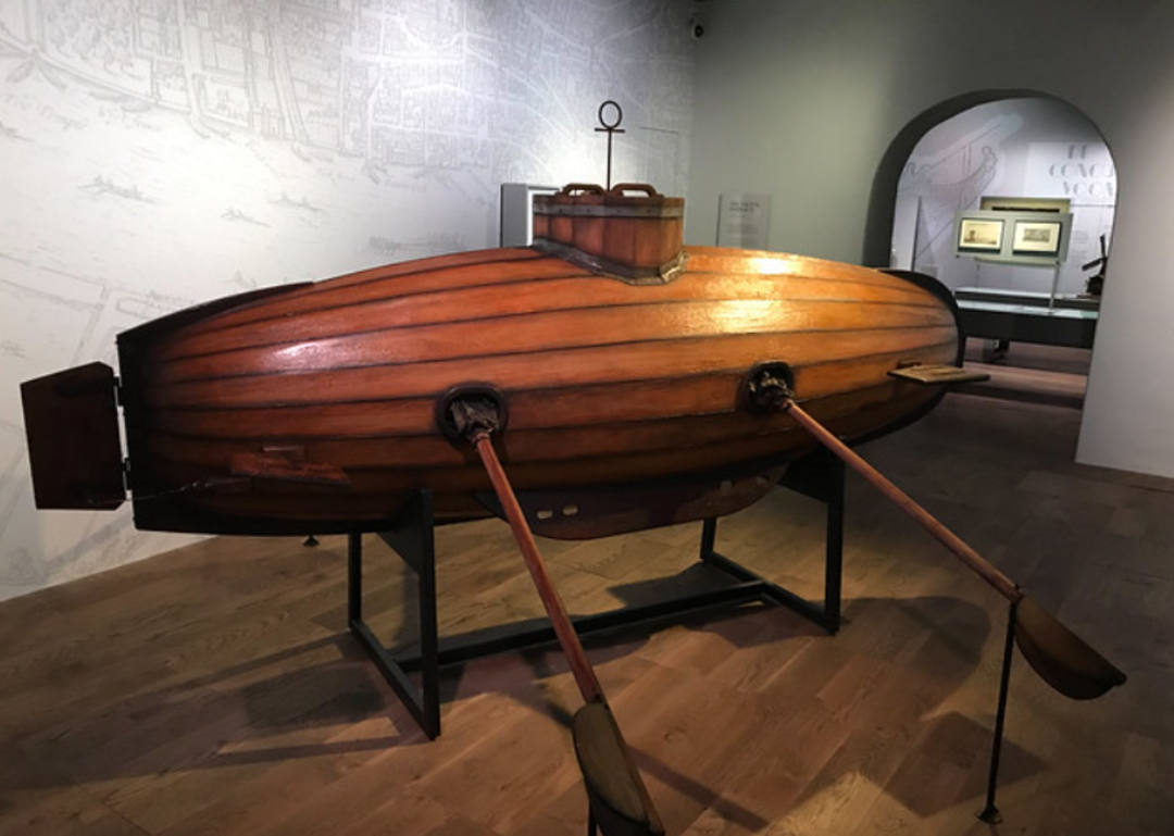 A wooden, oval-shaped early submarine with two oars, displayed on a stand in a museum room with wooden floors and historical maps on the walls.