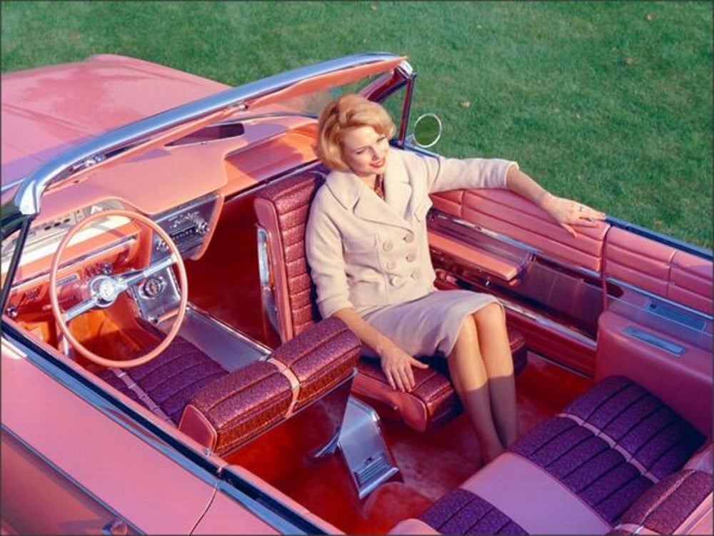 A woman in a light coat and dress sits in a vintage pink convertible car with matching pink and purple upholstery, parked on grass. She rests her arm on the back seat and looks thoughtfully into the distance.