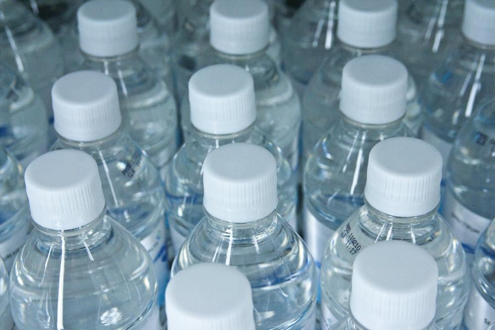 Rows of clear plastic water bottles with white caps, closely packed together, viewed from above. The bottles are filled with water and have labels on them.