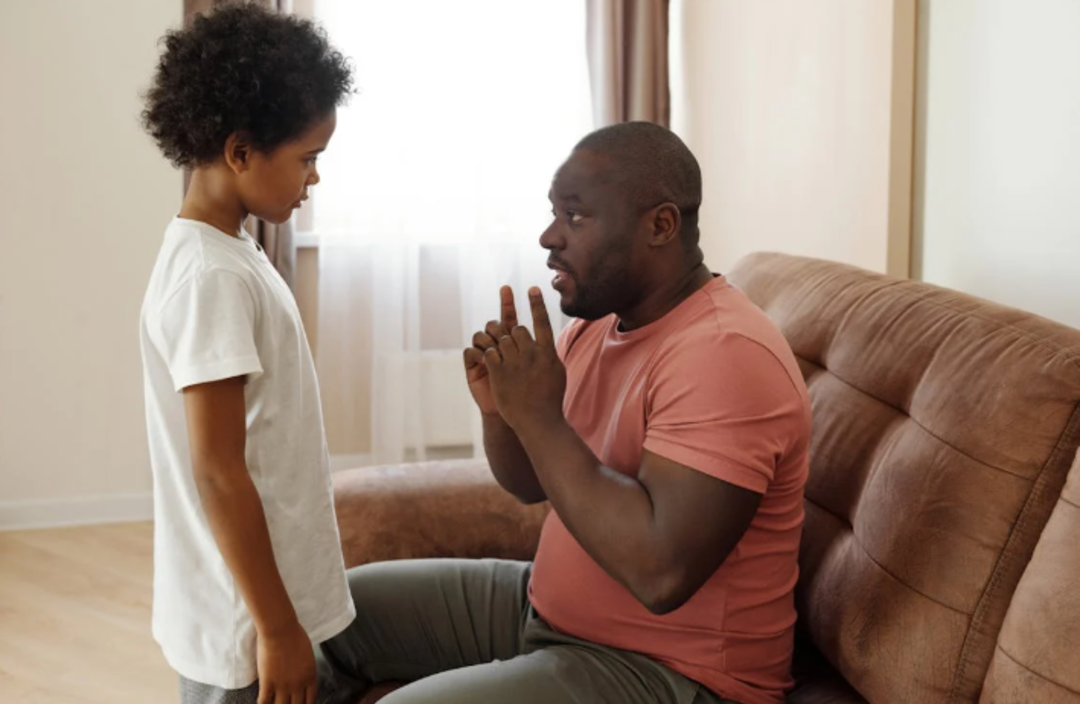 A man sitting on a couch holds up two fingers while talking seriously to a young boy standing in front of him. The boy looks down, listening intently. They are indoors with light coming through the window.