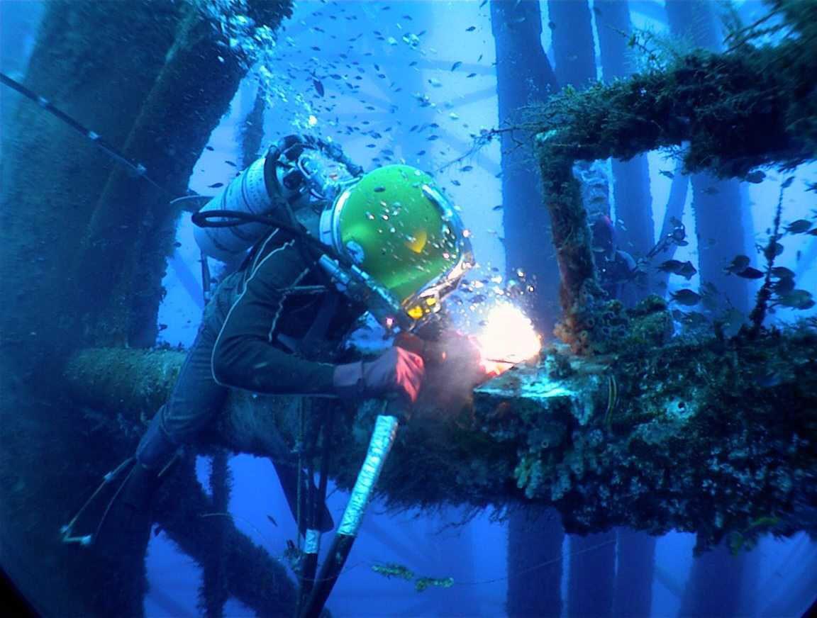 A diver in a yellow helmet and wetsuit performs underwater welding on a metal structure, surrounded by fish and beams covered in marine growth in deep blue water.