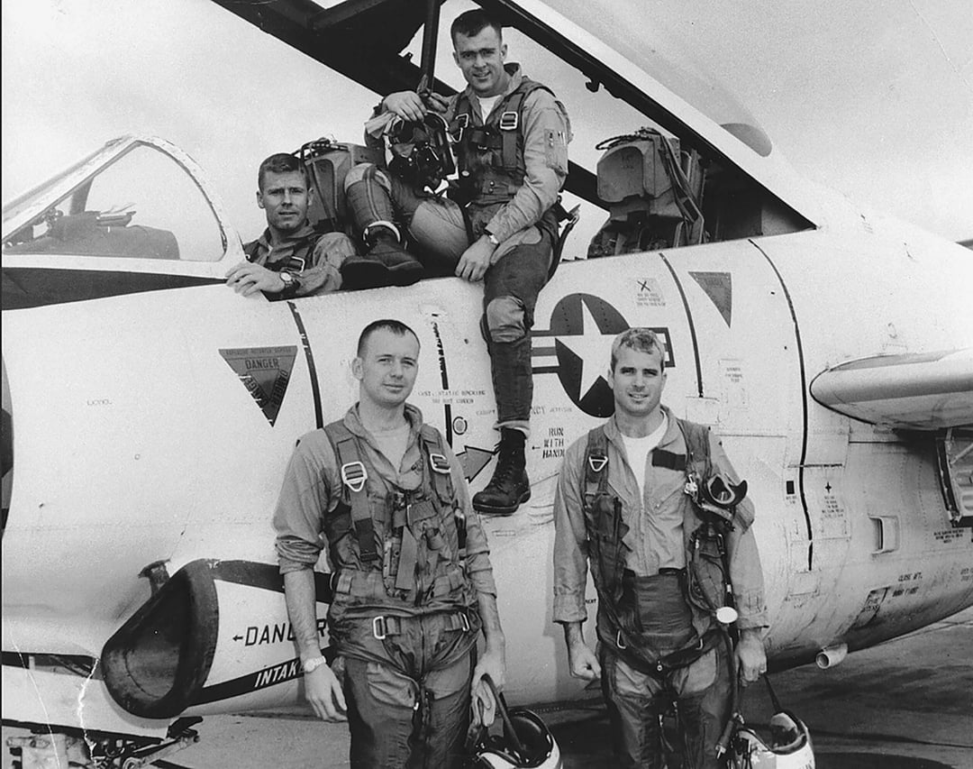 Four pilots in flight suits pose by a military jet, two standing on the ground and two sitting on the plane. The aircraft has visible U.S. Navy markings. The photo is in black and white.