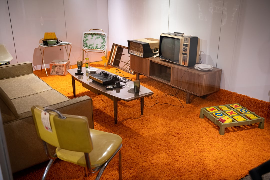 A retro living room with orange shag carpet, a vintage TV on a wooden stand, an old sofa, a coffee table with electronics, a yellow chair, and a tic-tac-toe game on the floor.