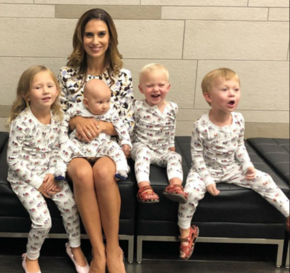 A woman sits on a bench with four young children. Three of the children and the baby on her lap wear matching pajamas with small prints. The woman smiles, while the children display various playful expressions.