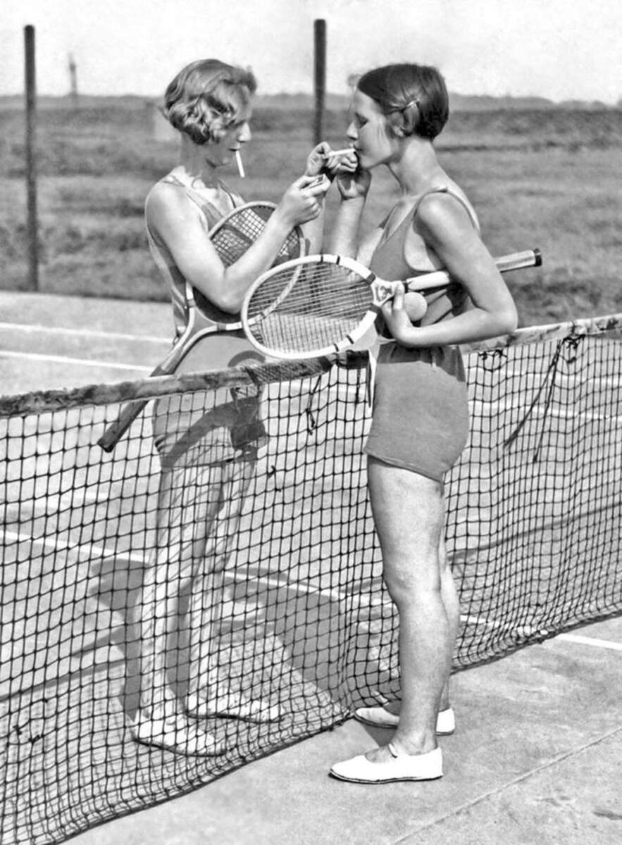 Two women in vintage tennis outfits stand on opposite sides of a net; one holds tennis rackets and balls while lighting a cigarette for the other, who is already smoking. The background shows an outdoor tennis court.