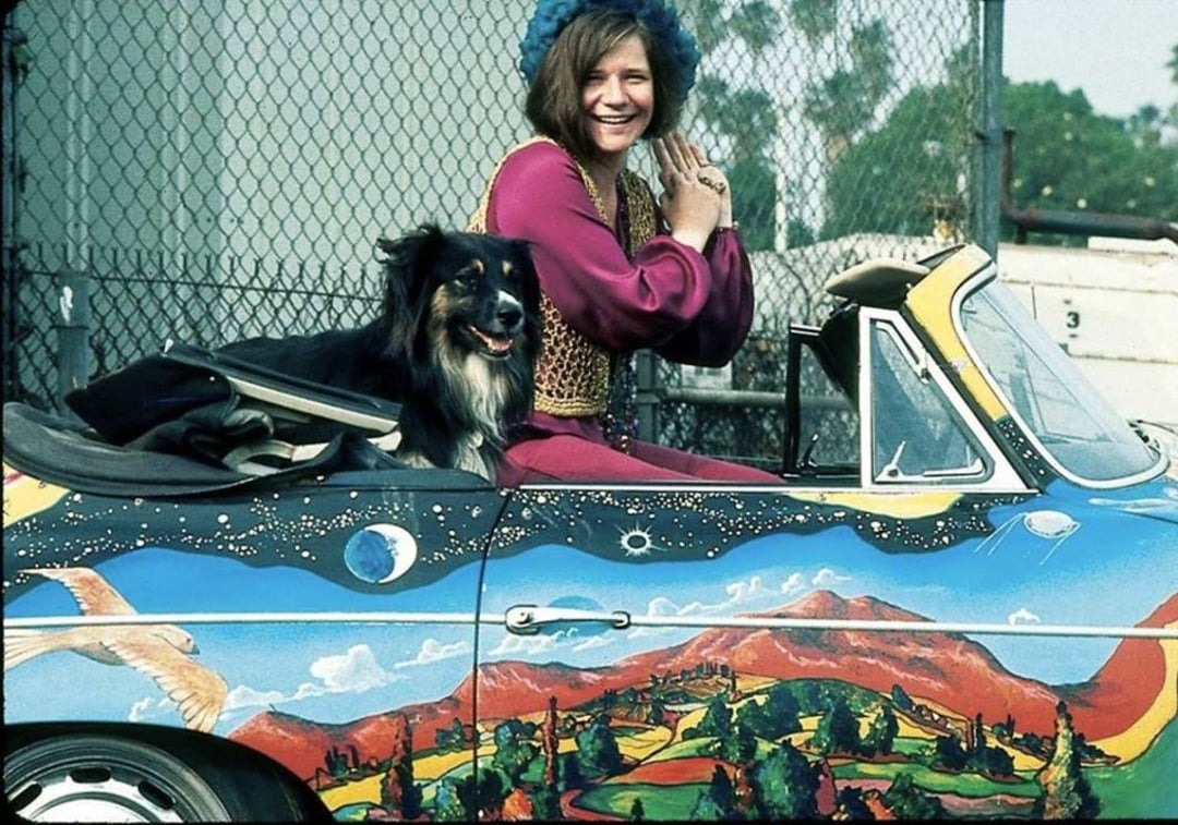 A smiling woman in colorful clothing and a blue hat sits in a convertible with a black dog. The car is painted with a vibrant, psychedelic landscape featuring mountains, birds, and planets. A chain-link fence is in the background.