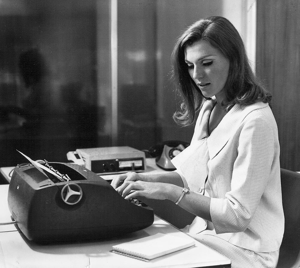 A woman in professional attire sits at a desk, typing on a vintage typewriter. There is a notepad in front of her and office equipment in the background.