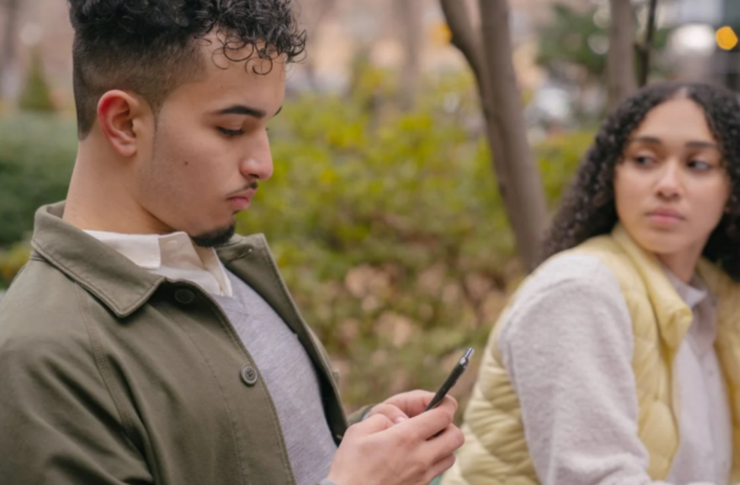 A young man looks at his phone while a young woman sitting beside him glances at him with a concerned expression. They are outdoors on a bench, surrounded by greenery.