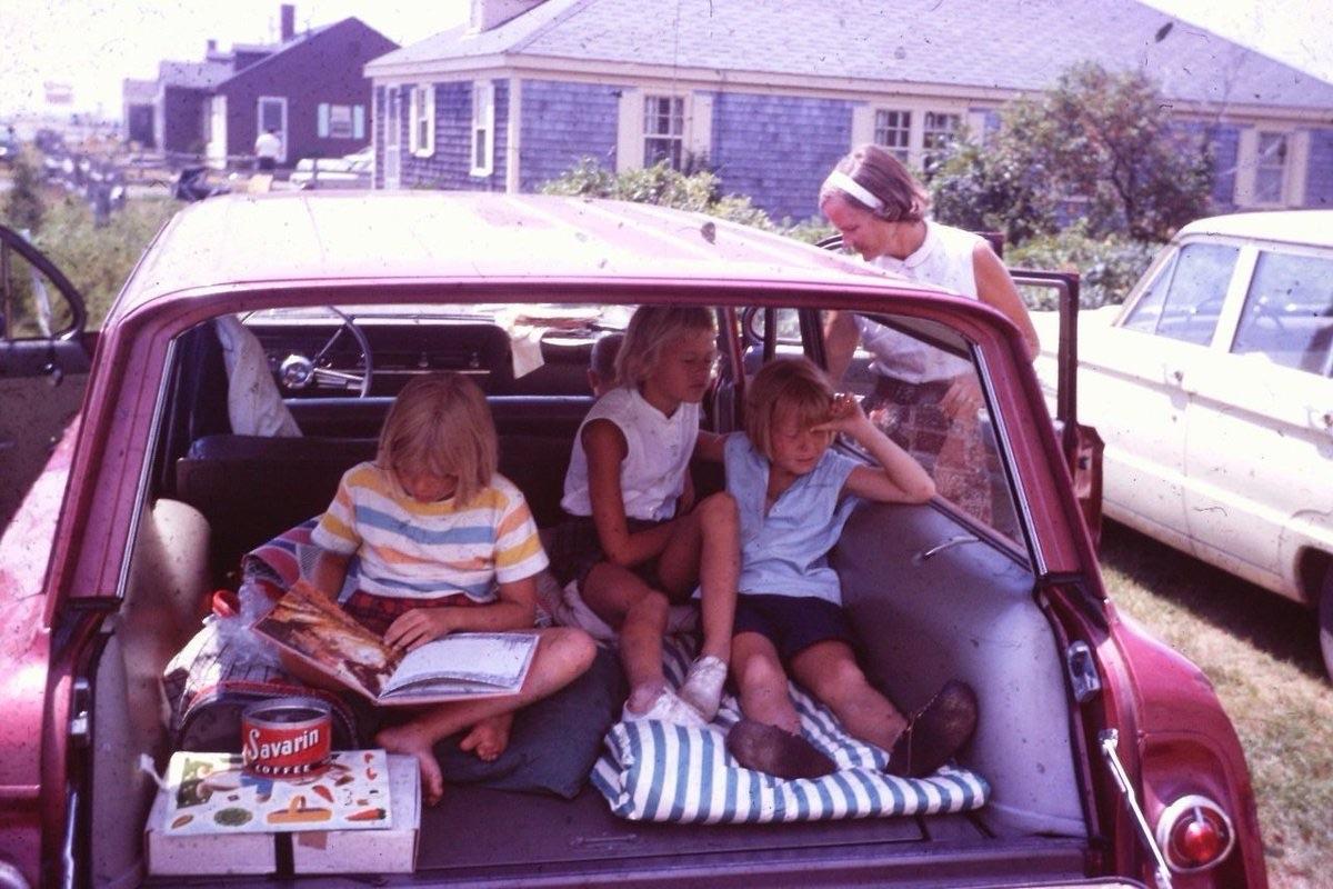 Three children sit in the open back of a red vintage station wagon, reading and playing. An adult woman stands nearby. A house and another car are visible in the background on a sunny day.