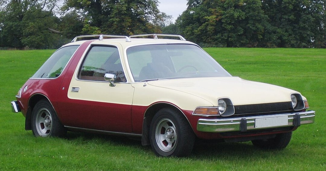 A vintage two-tone car with a cream upper body and red lower body is parked on grass, surrounded by trees. The car has a rounded design, roof rack, and large windows.
