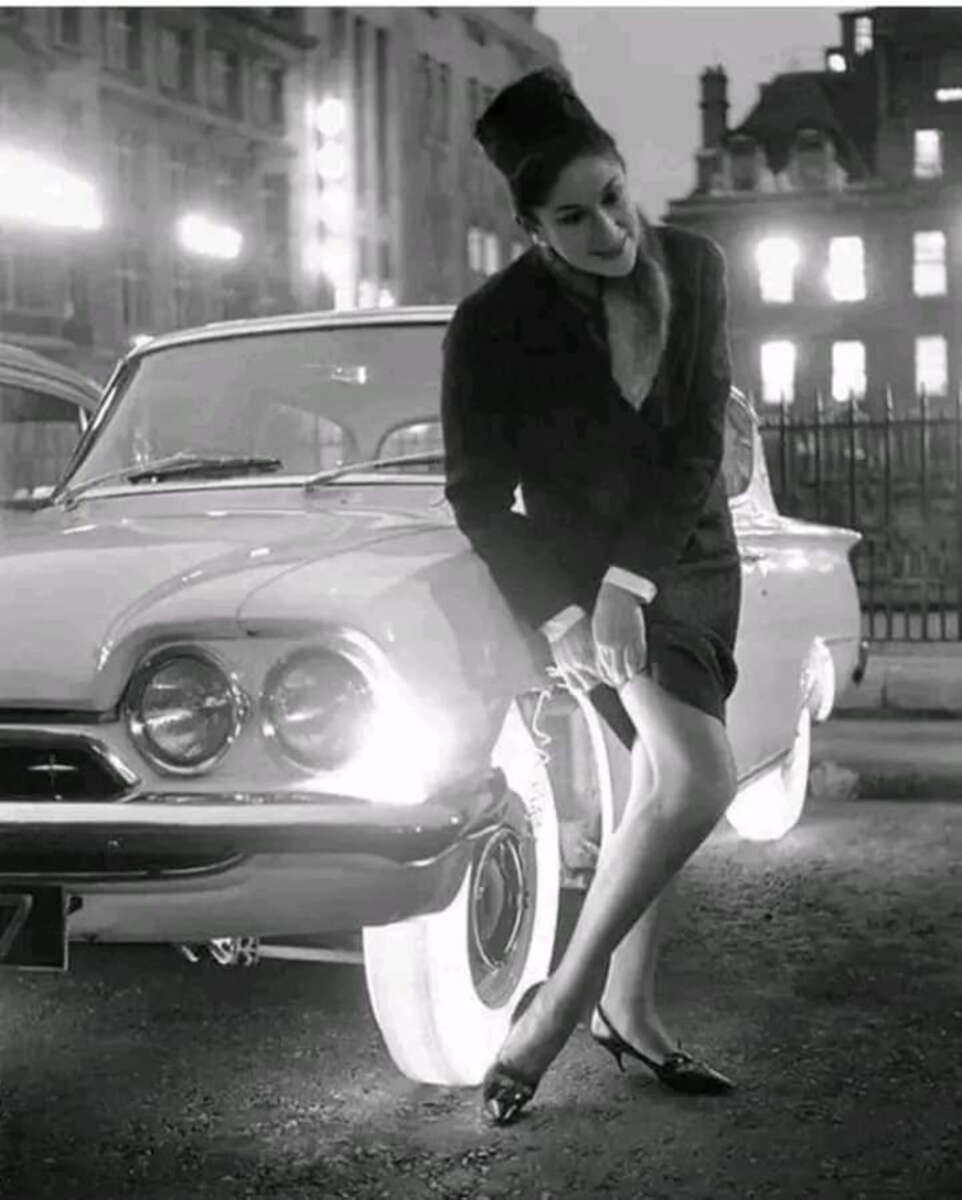 A woman in a dark dress and heels leans against a vintage car with glowing whitewall tires at night on a city street, with historic buildings in the background. The scene is in black and white.