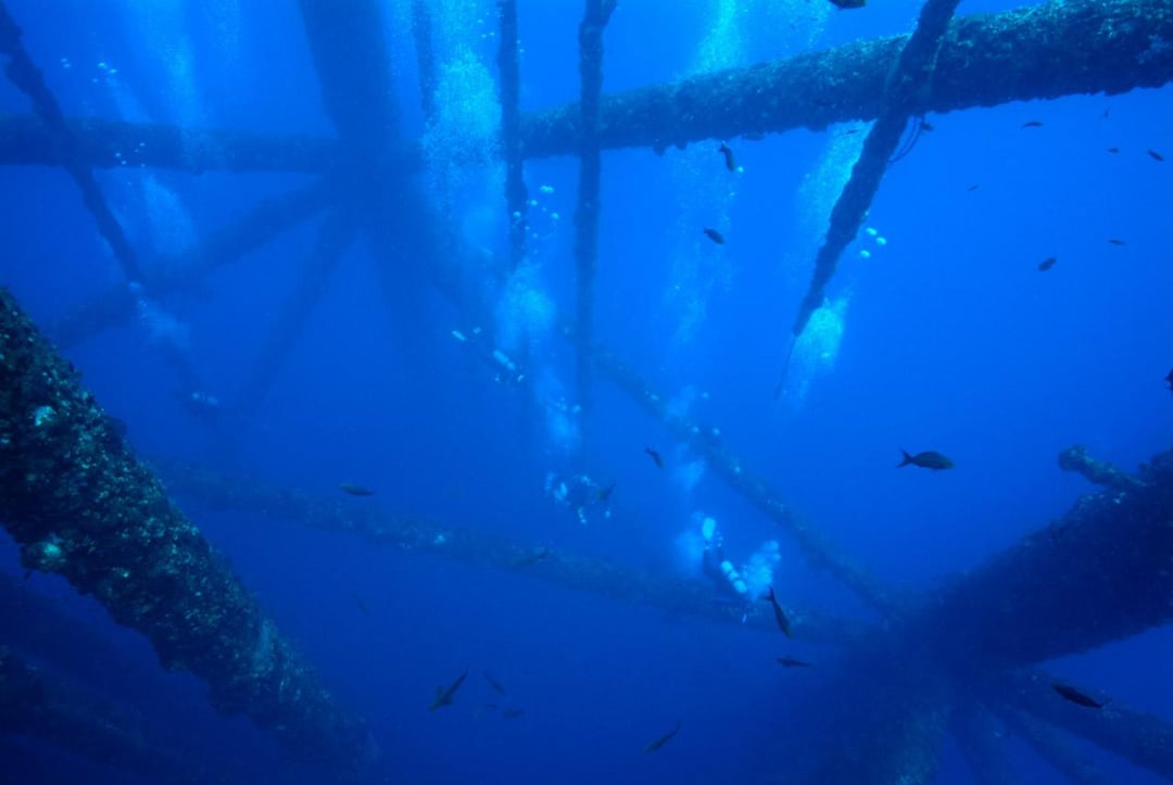Underwater view of divers ascending among the metal beams of an offshore oil platform, surrounded by small fish and rising bubbles in deep blue water.