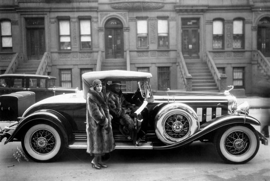 A Black man and woman in fur coats stand by a luxurious vintage car parked on a city street lined with brownstone buildings and stoops, evoking a 1920s or 1930s urban scene.