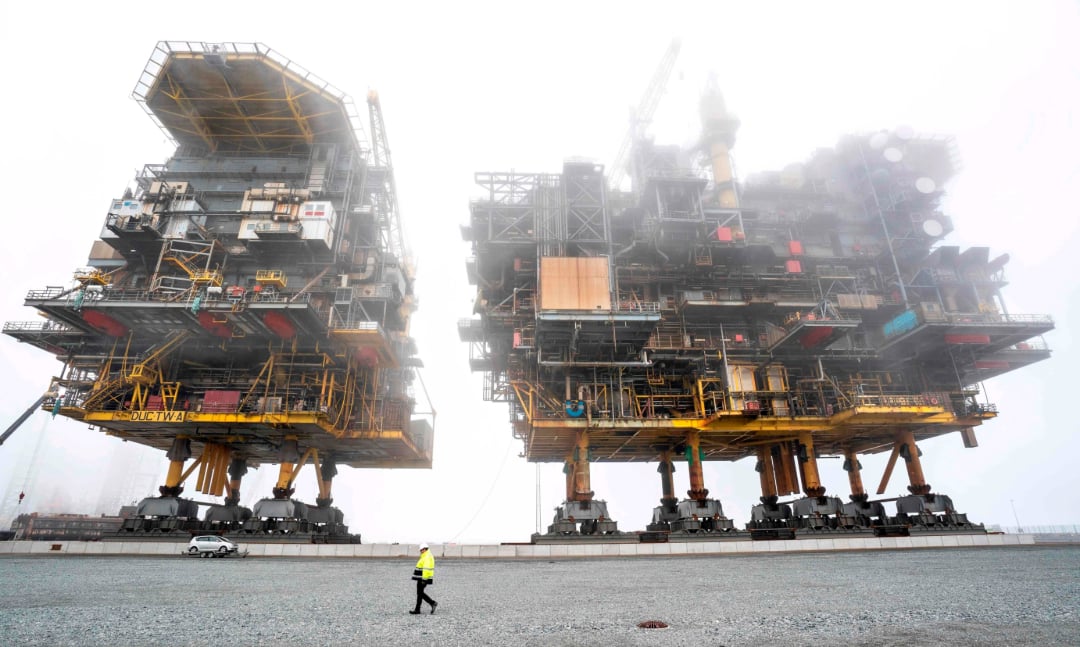 Two massive offshore oil platform structures stand on land amid fog, with a small car and a person in a yellow safety jacket walking in the foreground, highlighting the immense scale of the platforms.