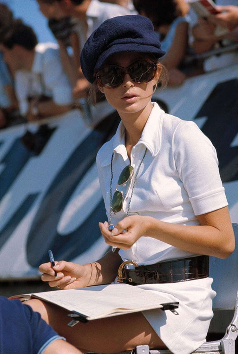 A woman in a white dress, dark sunglasses, and a navy cap sits outdoors holding a pen and clipboard. She wears a large belt and has a pair of sunglasses hanging from her neck. A blurred crowd is visible in the background.