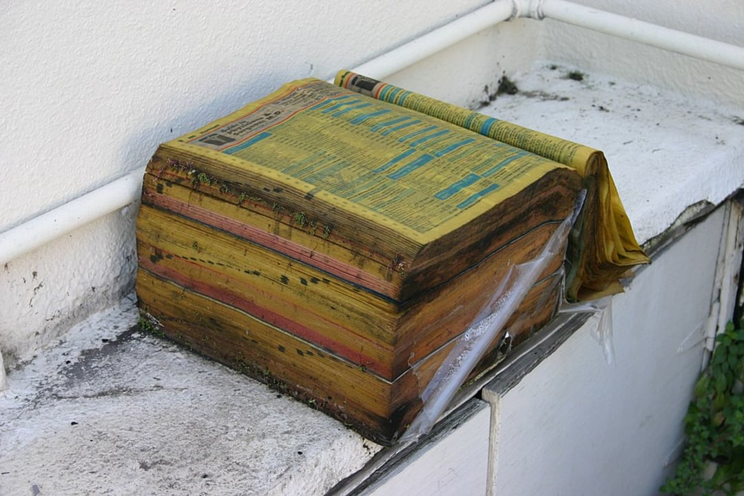 A thick, weathered phone book with yellowed, curled pages sits outdoors on a concrete ledge, partially wrapped in torn plastic, showing signs of age and exposure to the elements.