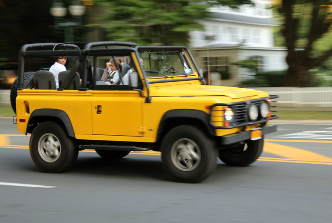 A yellow convertible SUV with three people inside drives down a street lined with trees and a white house in the background. The vehicle and passengers appear in motion, with the background blurred.