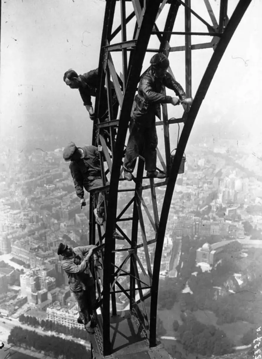 Four workers climb and perform maintenance high on the steel structure of the Eiffel Tower, with a view of Paris and its buildings far below in the background.