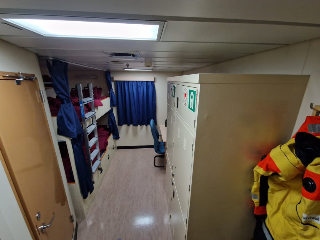 A narrow cabin room with bunk beds on the left, blue curtains, lockers on the right, a desk with a chair near the window, and yellow survival suits hanging on hooks by the door.