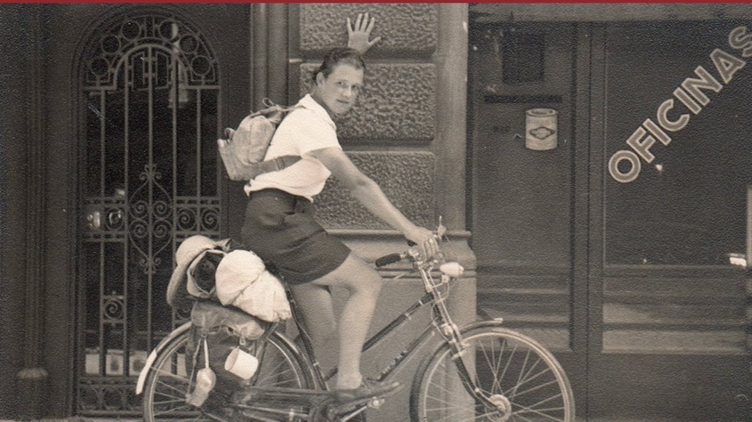 A person wearing a skirt and backpack sits on a bicycle, one hand on the handlebars and the other touching a wall. Laundry or bags are attached to the back of the bike. The background shows a building with the word "OFICINAS.