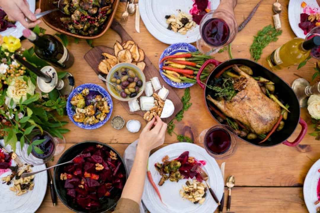 A table set for a meal with roasted meat, vegetables, salads, bread, olives, cheese, wine, and flowers. Several hands are reaching for food, creating a lively, communal dining scene.