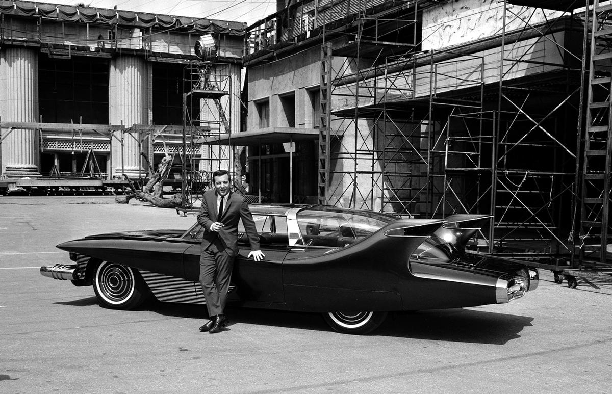 A man in a suit leans against a futuristic, sleek car with dramatic fins, parked in front of a building under construction with scaffolding and columns visible in the background.