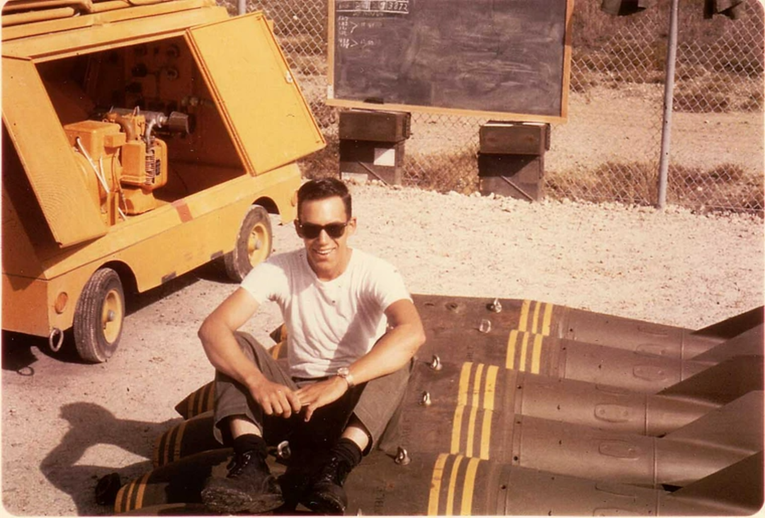 A man wearing sunglasses and a white t-shirt sits on the ground beside several large bombs with yellow stripes. There's a yellow generator and a chalkboard on stands behind him, all within a fenced outdoor area.