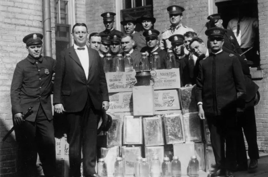 A group of police officers and men pose with confiscated boxes and glass jars, likely containing illegal alcohol, stacked against a brick building during the Prohibition era.
