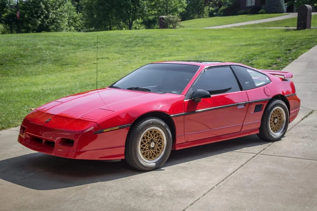 A red classic sports car with pop-up headlights and gold mesh wheels is parked on a driveway, with green grass and trees in the background on a sunny day.