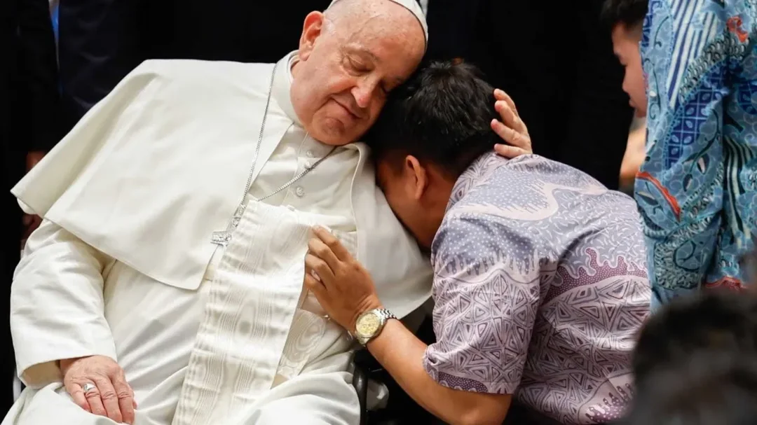 A person in a patterned shirt embraces and leans their head on an elderly man in white religious attire and a cap, who gently holds them and closes his eyes, surrounded by others.