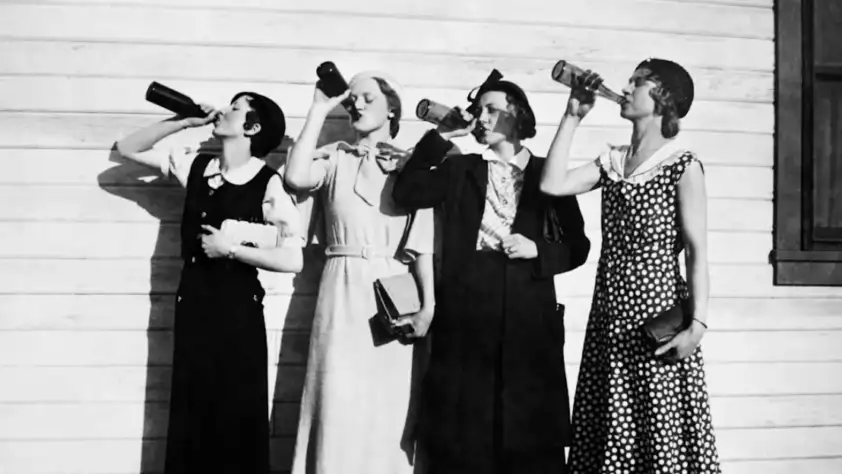 Four women in vintage dresses stand side by side against a wooden wall, each drinking from a bottle and holding books or purses, in a black and white photo.